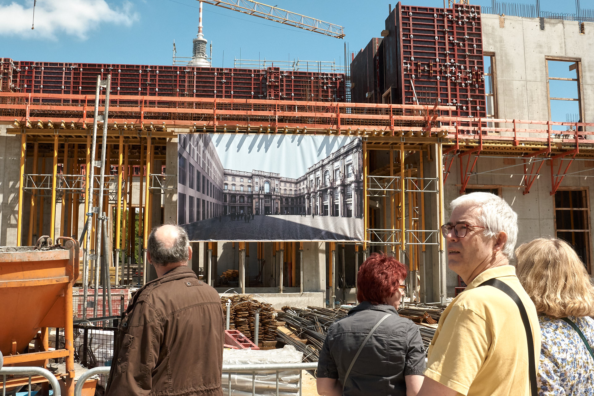 Menschen betrachten die Baustelle des Humboldtforums, vor ihnen Bretter und Stahlrohre, ein Baugerüst, an dem ein Bild des zukünftigen Schlüterhofes hängt.